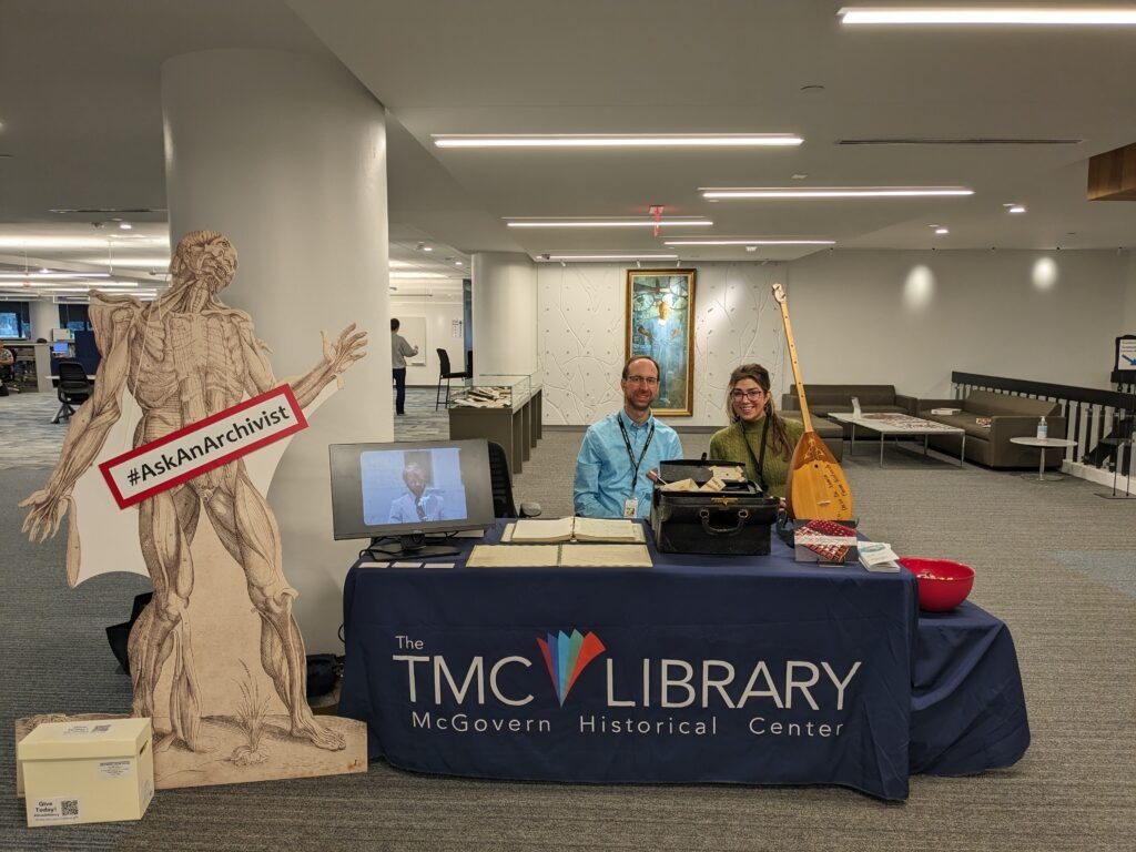 Matt Richardson and Tara Sims welcome questions at the Ask an Archivist Booth in the TMC Library in October 2023. They sit behind a table exhibiting collection items. Table cloth reads The TMC Library, McGovern Historical Center.