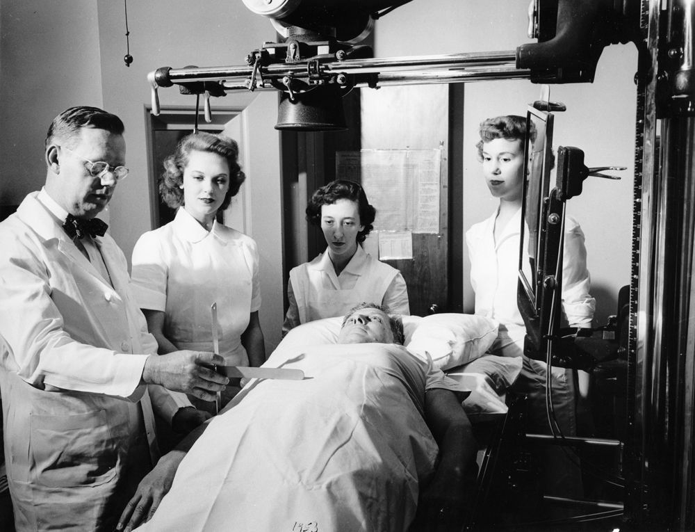 Radiology Department X-ray Technicians are trained by Dr. Luther M. Vaughn, 1953. From left to right: Dr. Luther M. Vaughn, Nancy Rogers, Suzi MacAllister, and Margaret Echols, playing the patient is Walter Sterling. [McGovern Historical Center, Gift of Dr. Luther M. Vaughn, Photo Files, Institutions and Organizations, Hermann Hospital Radiology Department, 1953]