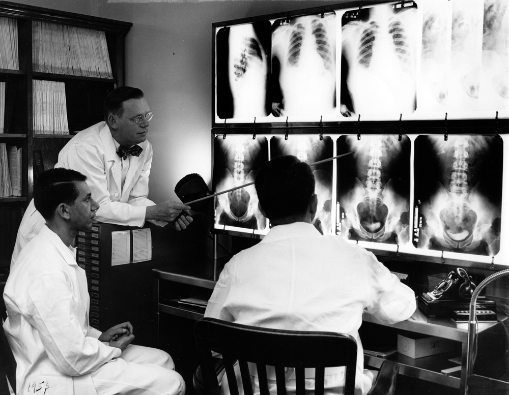 Examining X-ray photographs at Hermann Hospital, 1953. Seated at left is a resident from Iraq, Dr. Mohamed Aba Tabik (Dr. Mo), standing is Dr. Luther Vaughn, seated facing the light stand is Dr. William Owsley. [McGovern Historical Center, Gift of Dr. Luther M. Vaughn, Photo Files, Medical Equipment and Apparatus, Hermann Hospital X-ray equipment, 1953]
