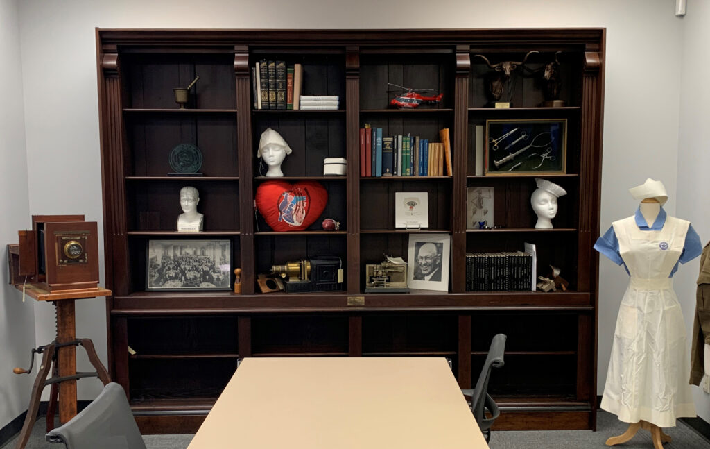 [Oliver Wendell Holmes, MD, bookcases in the McGovern Historical Center reading room. The camera to the left of the bookcases and the magic lantern on the bottom shelf were donated by Detering.]